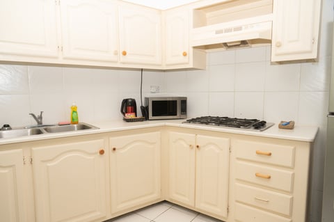 Small cream-colored kitchen with white subway tile backsplash, stovetop, microwave, and sink with cabinets and wooden handles