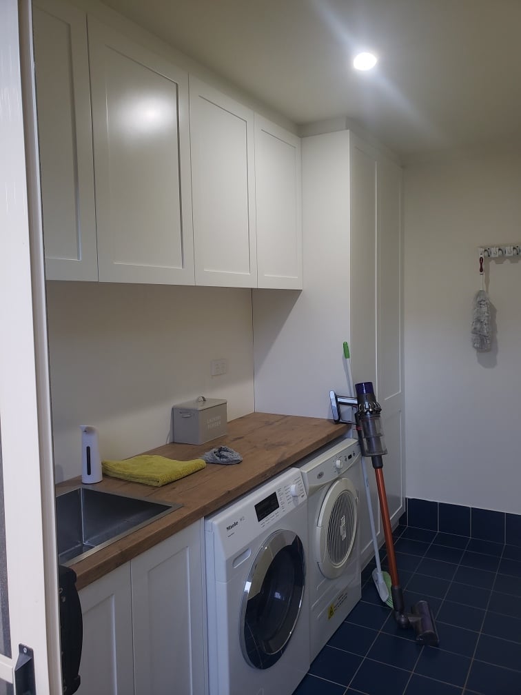 Modern laundry room with white cabinetry, two front-load washing machines, wooden countertop, and dark tile flooring