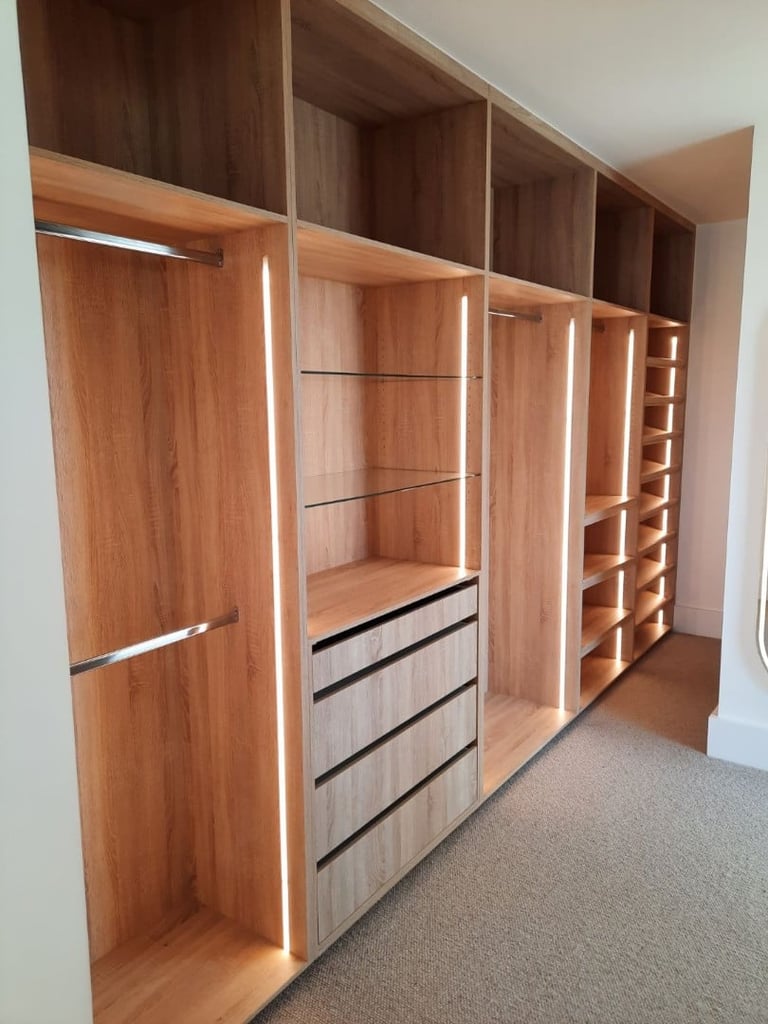 Custom wooden walk-in closet with multiple compartments, shelving, drawers, and glass doors with modern lighting