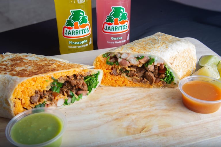 Two filled tacos with cheese and meat on a wooden board, served with green and red salsas and Jarritos beverages