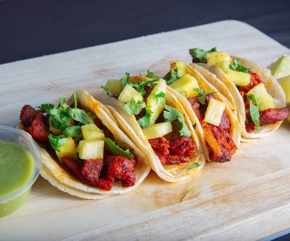 Four al pastor tacos with pineapple and cilantro on a wooden board