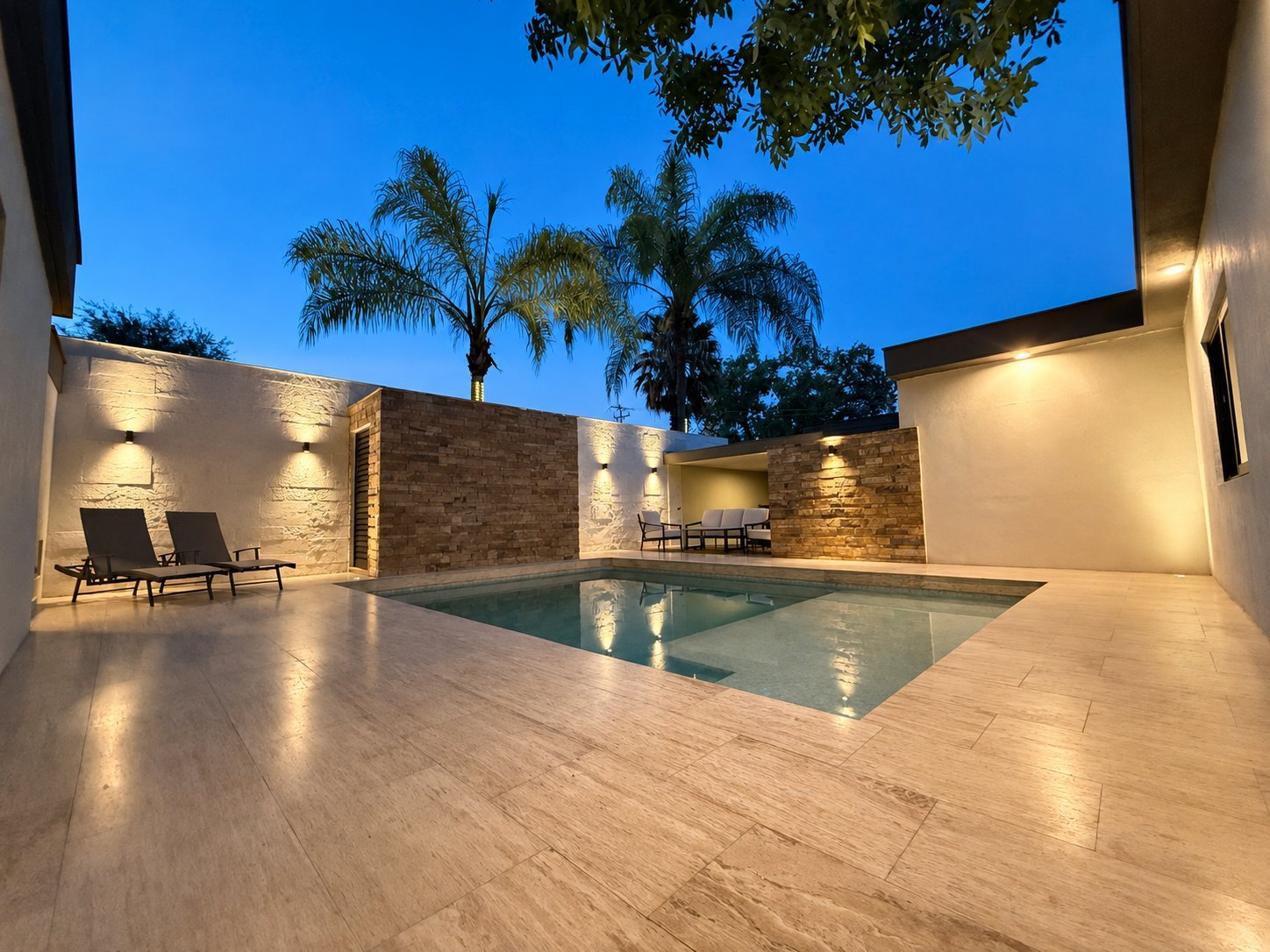 Modern residential courtyard with rectangular swimming pool, lounge chairs, palm trees, and illuminated stone walls at dusk
