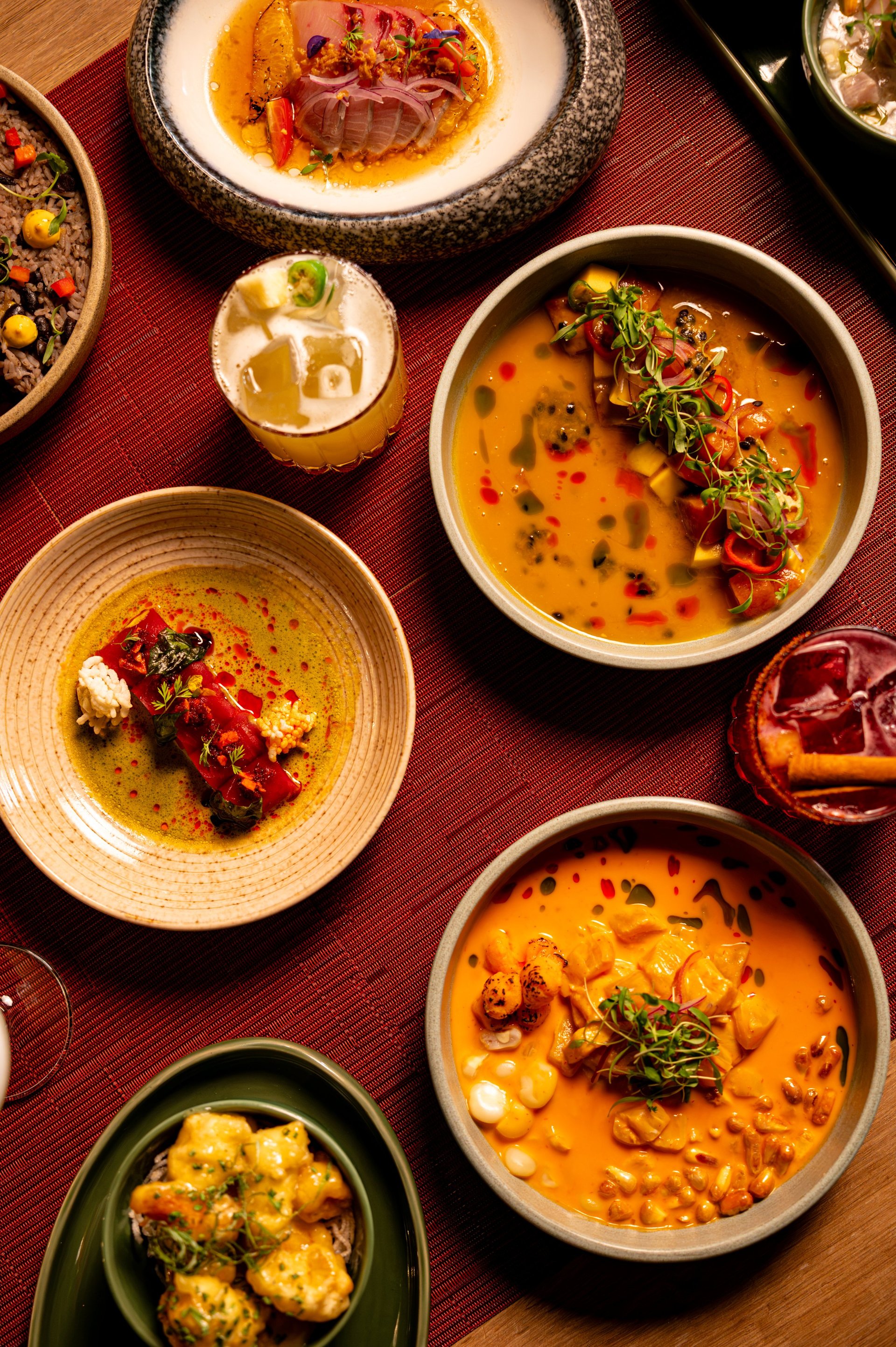 Overhead view of plated Indian appetizers and small dishes arranged on a burgundy tablecloth, featuring creamy soups, garnishes, and protein toppings