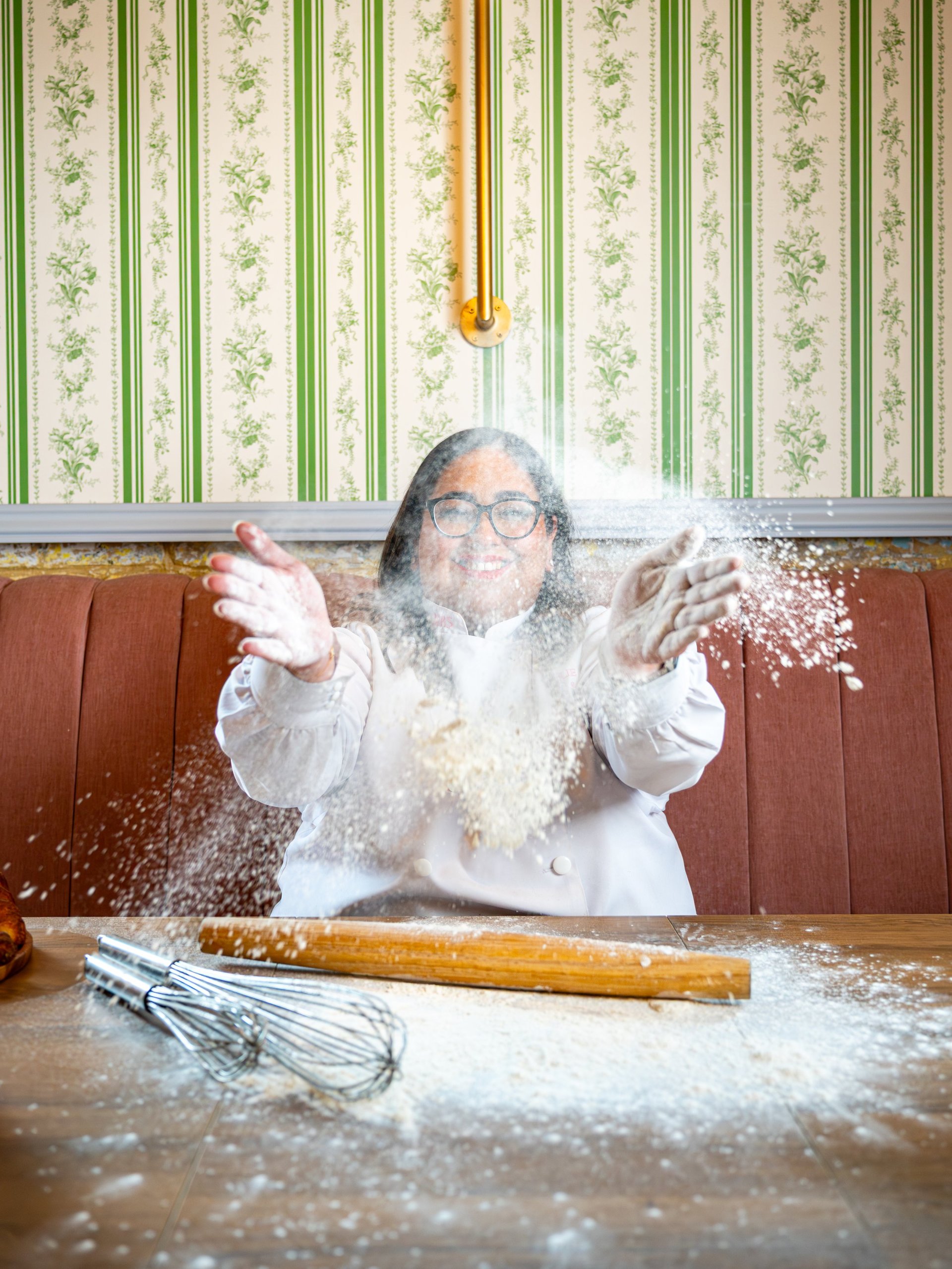 Person wearing white chefs coat and glasses dusted with flour, playfully tossing flour in the air at a kitchen counter with a rolling pin and whisk