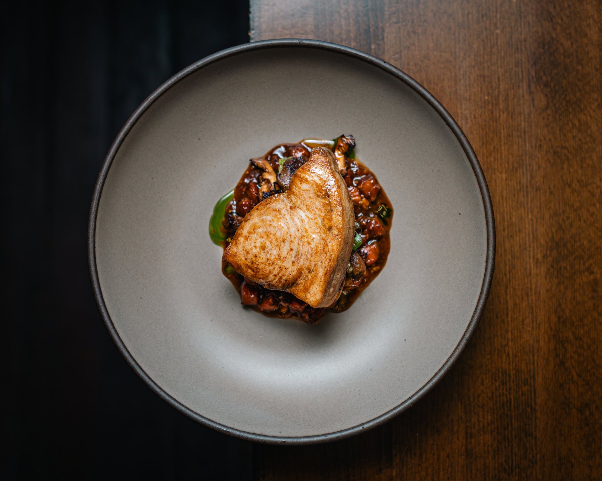 Seared scallop with dark caramelized sauce and basil leaf on a grey plate, overhead view