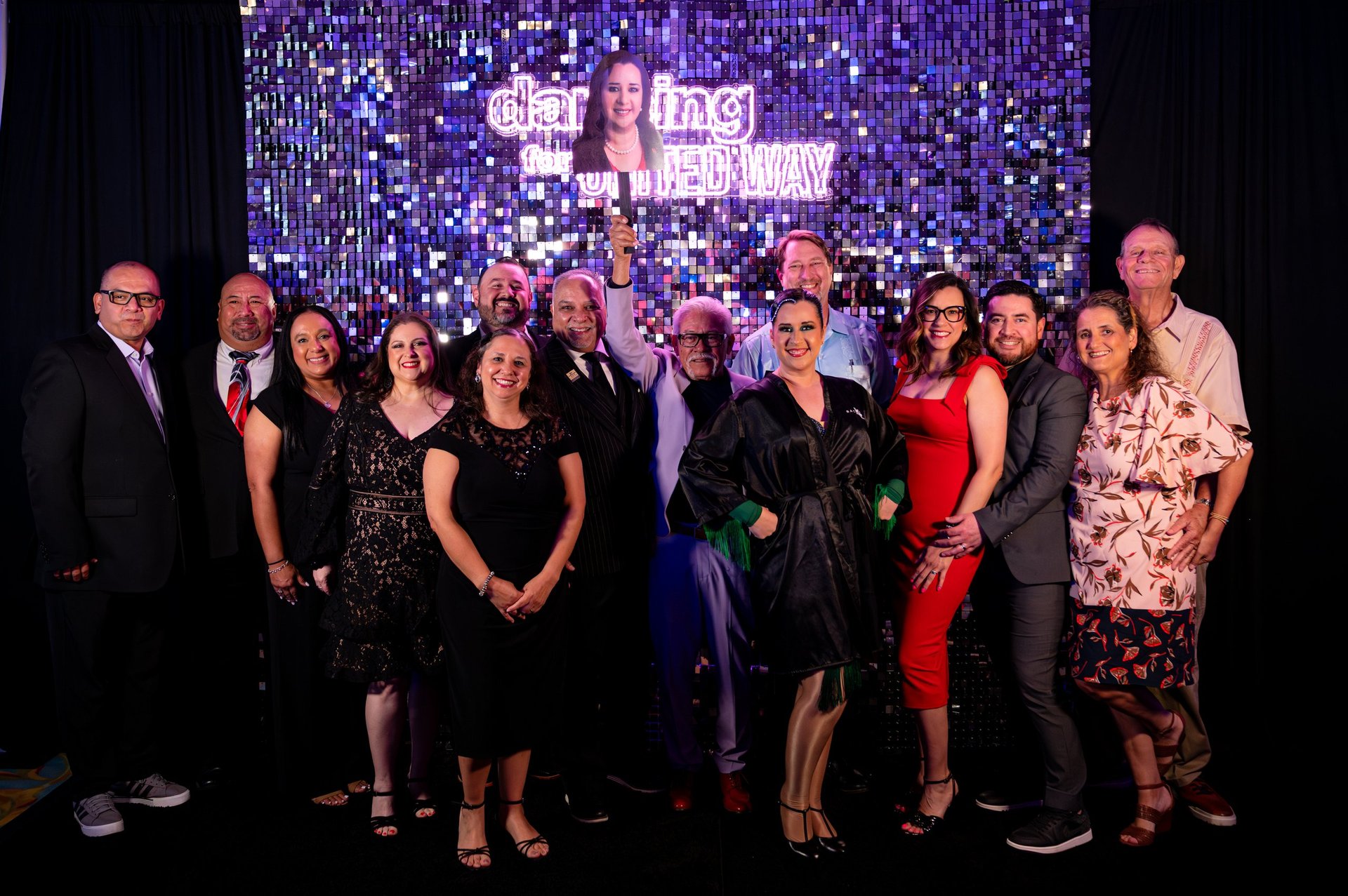 Group of people standing together on stage at an event with a purple sequined backdrop and text