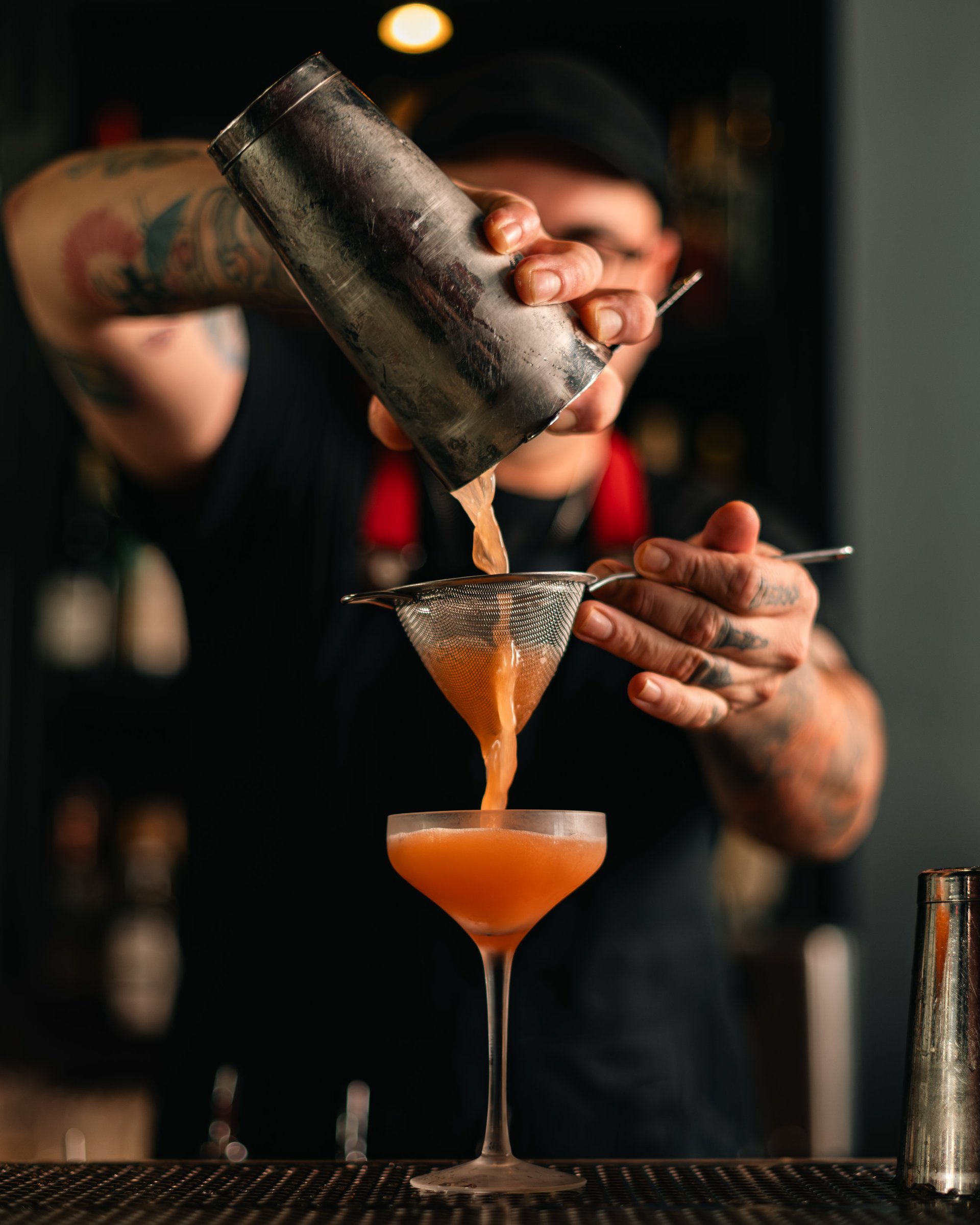 Bartender pouring cocktail from metal shaker into coupe glass, demonstrating mixology technique