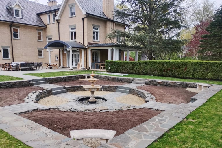 Large stone patio with circular fire pit in manicured backyard of elegant brick mansion with covered porch and hedges