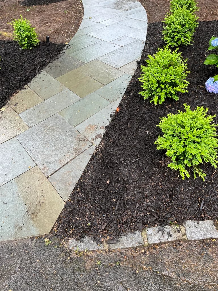 Curved stone pathway with dark mulch garden beds, bright green shrubs, and purple flowers alongside
