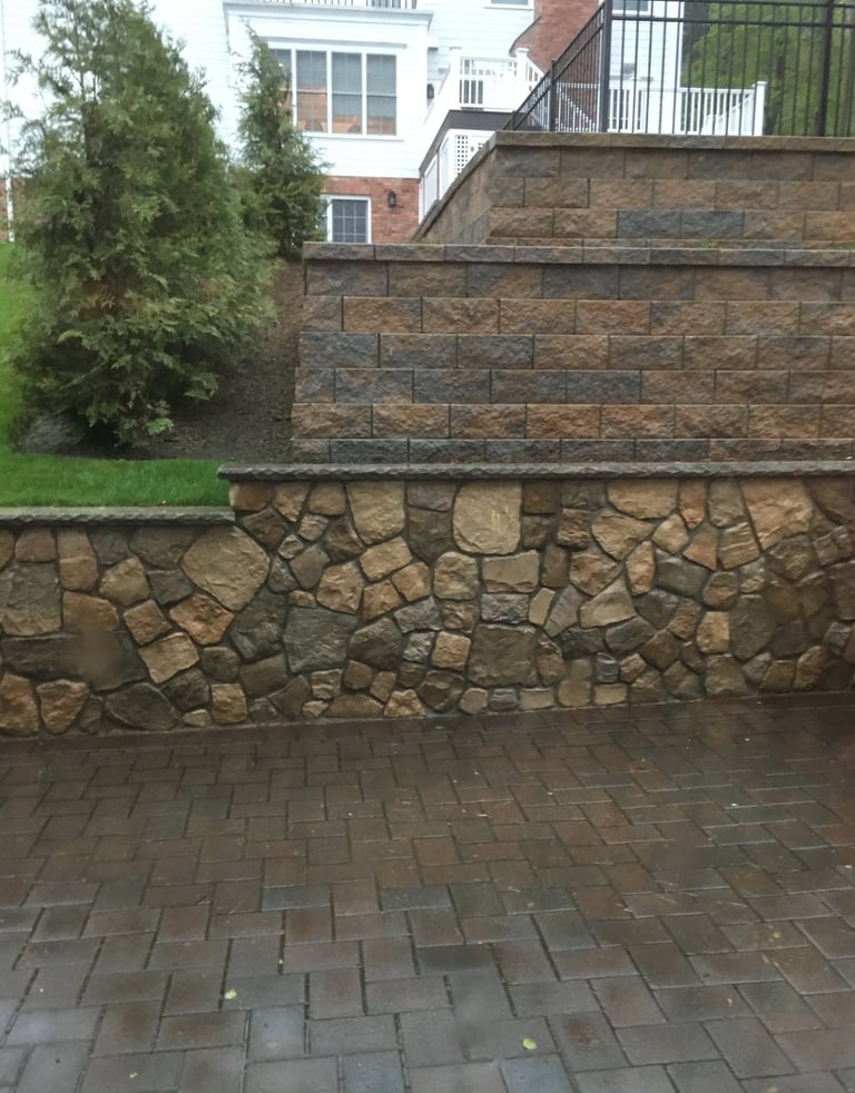 Stone and brick retaining wall with patio below, residential brick house and landscaping visible above