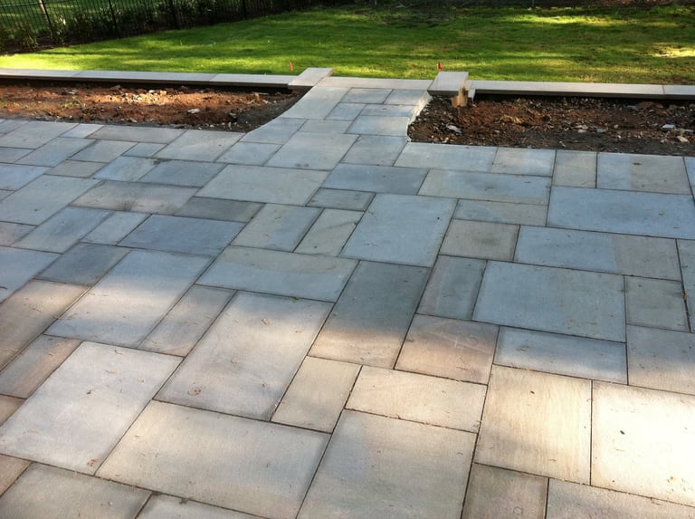 Paved patio with herringbone pattern stone tiles, adjacent to grass and mulch landscaping in sunlight