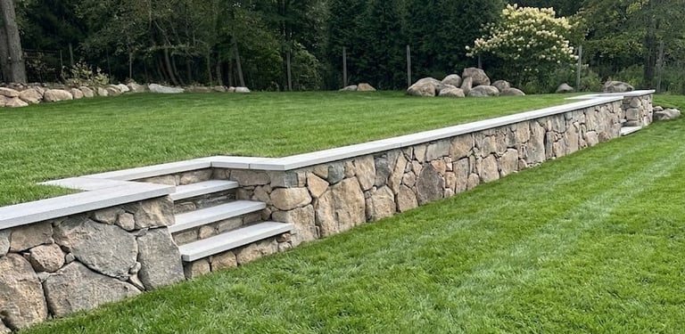 Long stone retaining wall with concrete cap and steps built on grassy lawn with forested background