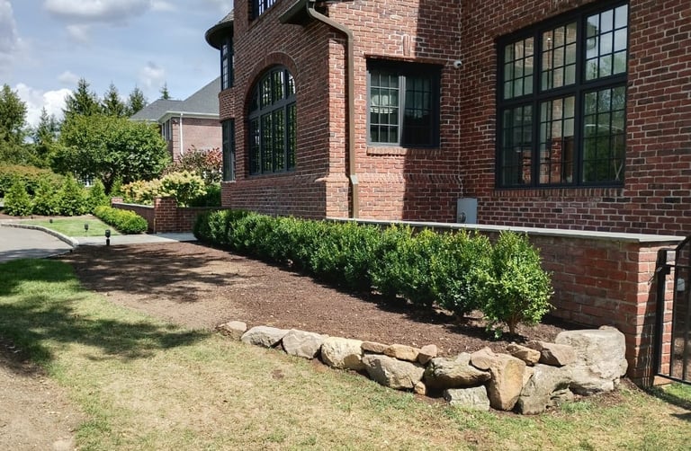 Brick house exterior with green windows, manicured landscaping, stone border, and lawn featuring boxwood shrubs