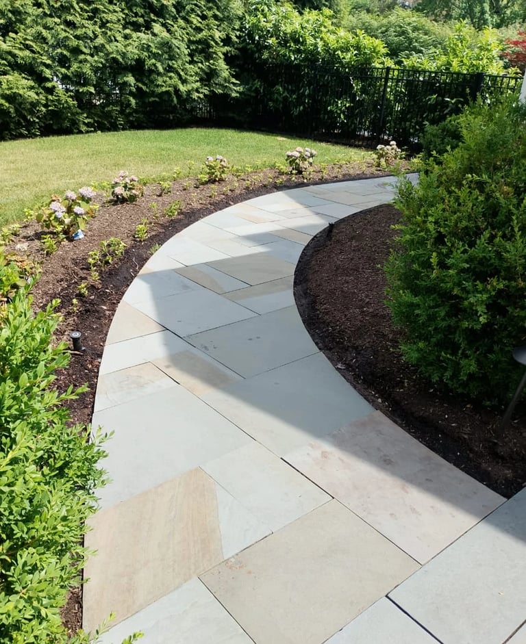 Curved concrete pathway winding through a manicured garden with landscaping beds, green lawn, and hedges