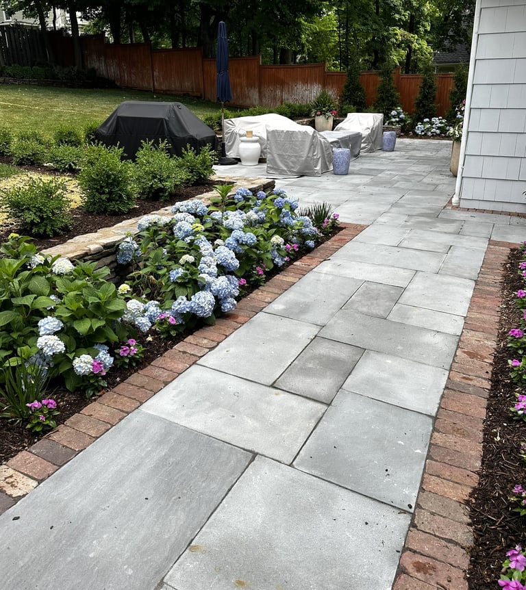 Backyard patio with stone pathway, blooming hydrangeas and pink flowers along brick borders, grass, wooden fence, and covered patio furniture