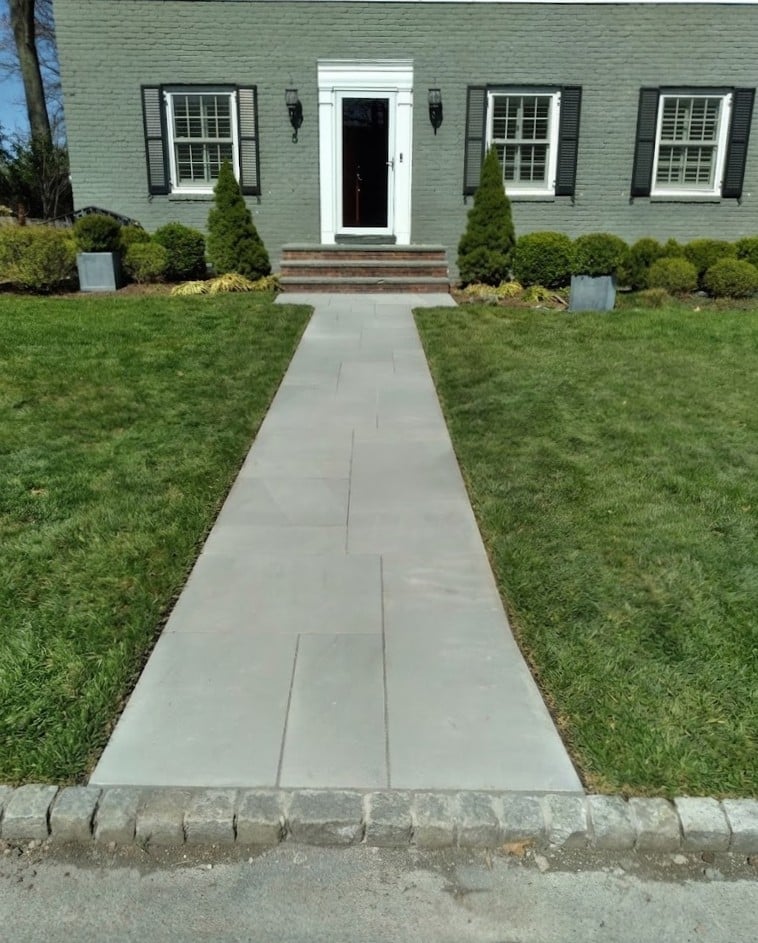 Pale green house with white door and windows, straight concrete walkway flanked by green lawn and foundation plantings leading to entrance