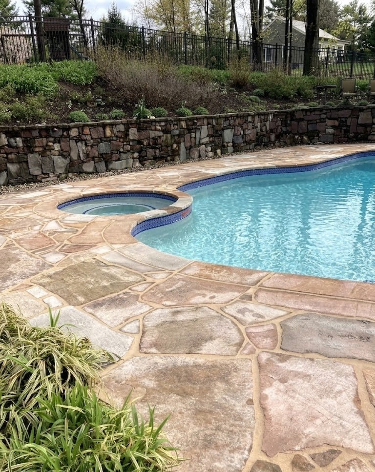 Residential swimming pool with spa featuring turquoise water, stone patio, and landscaped retaining wall with evergreen trees