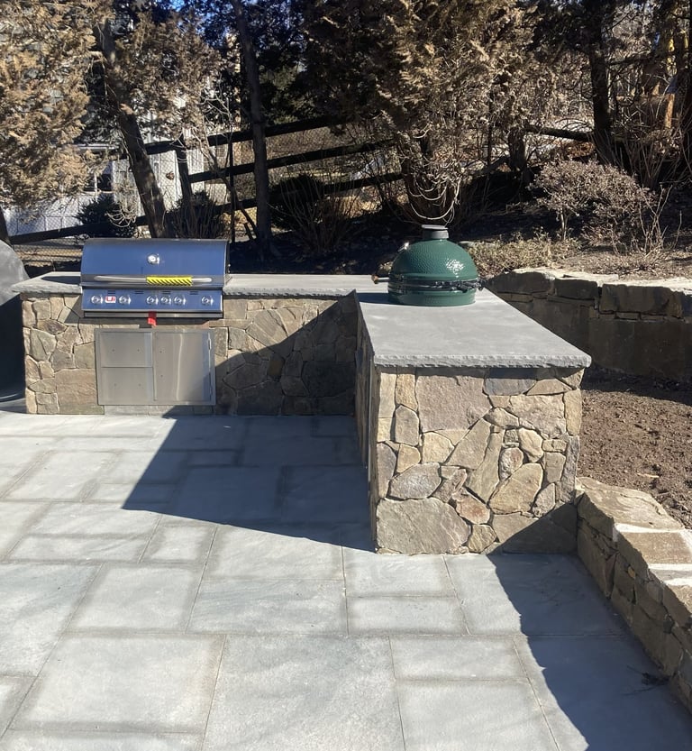 Outdoor kitchen setup with blue gas grill and green dome cooker on stone countertops in a patio area
