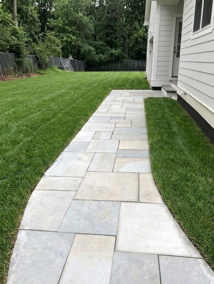 Stone pathway cutting through manicured green lawn beside white house with black fence and trees in background