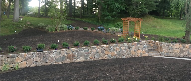 Garden bed with stone border, mulch, and wooden trellis surrounded by trees in landscaped yard