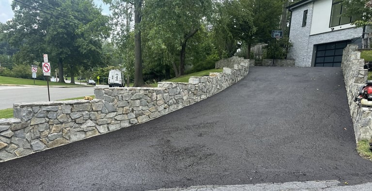 Asphalt driveway curving uphill past a stone retaining wall with trees and a house visible in the background