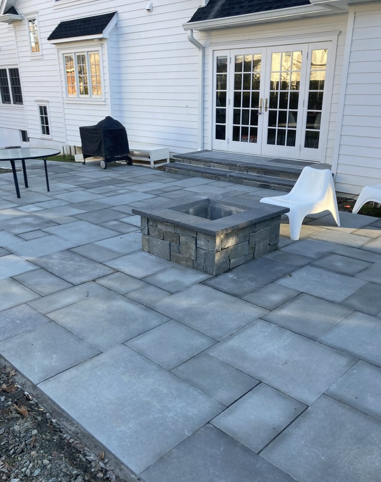 Backyard patio with gray stone pavers, rectangular fire pit, and white house with glass doors in background