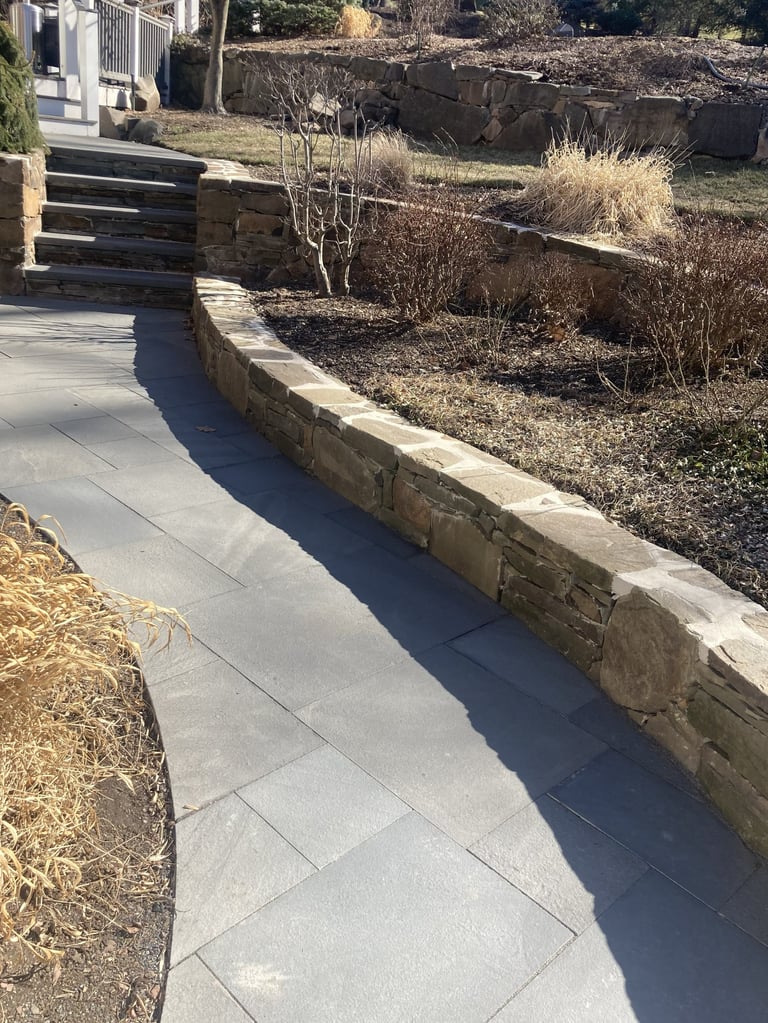 Concrete patio pathway with stone retaining wall bordered by dried plants and mulch, wooden steps visible in background