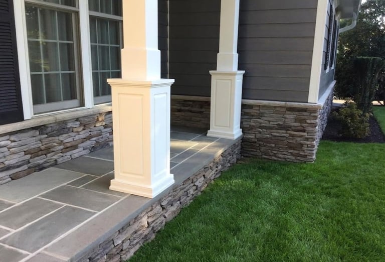 Front porch with white columns, stone base, large windows, and green lawn adjacent to the house exterior