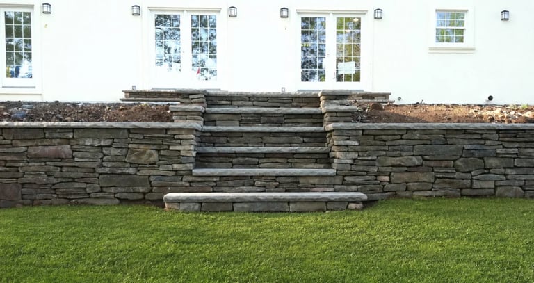 Slate stone retaining wall with centered steps leading up to a building with multiple windows and green lawn in foreground