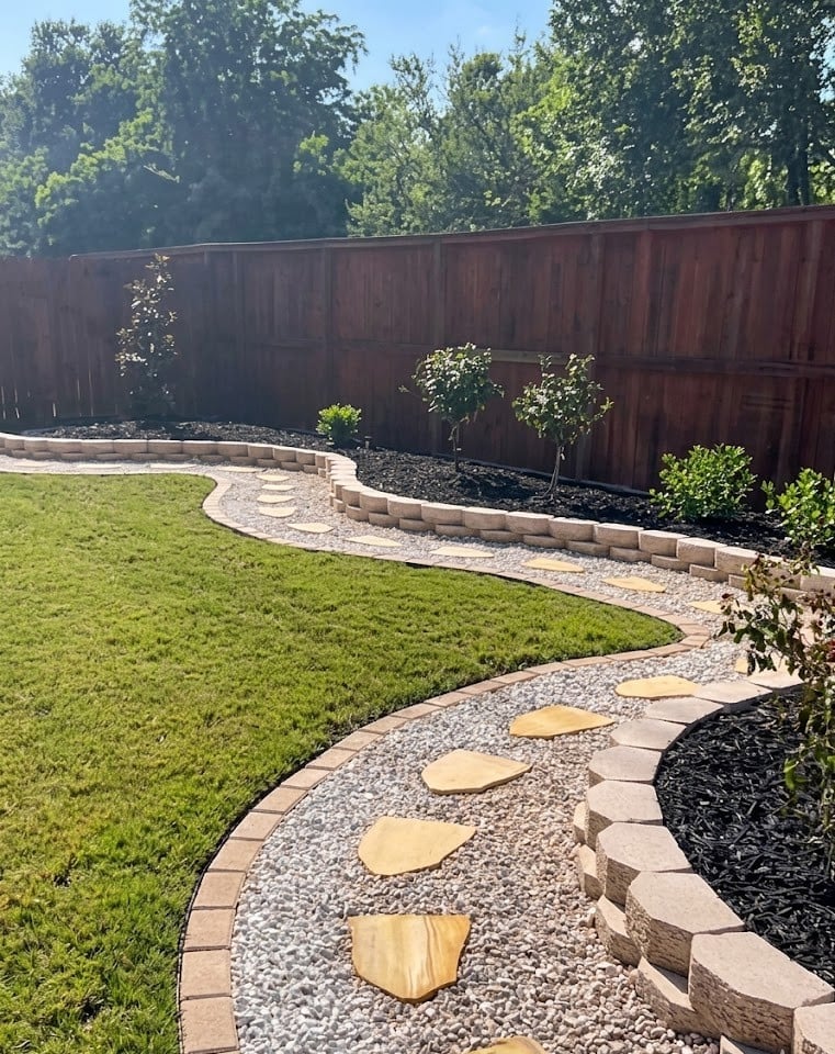 Backyard landscape with curved stone pathway, green lawn, wooden retaining wall, and lush trees