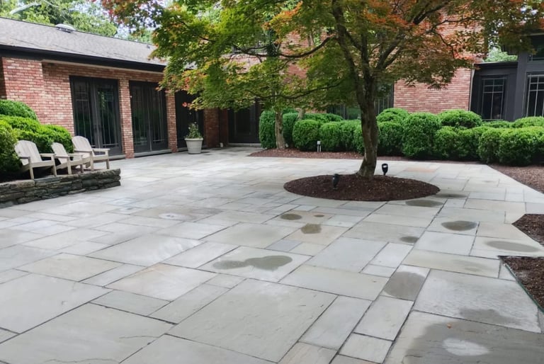 Modern patio courtyard with gray stone pavers, surrounded by red brick buildings, mature trees, and manicured hedges