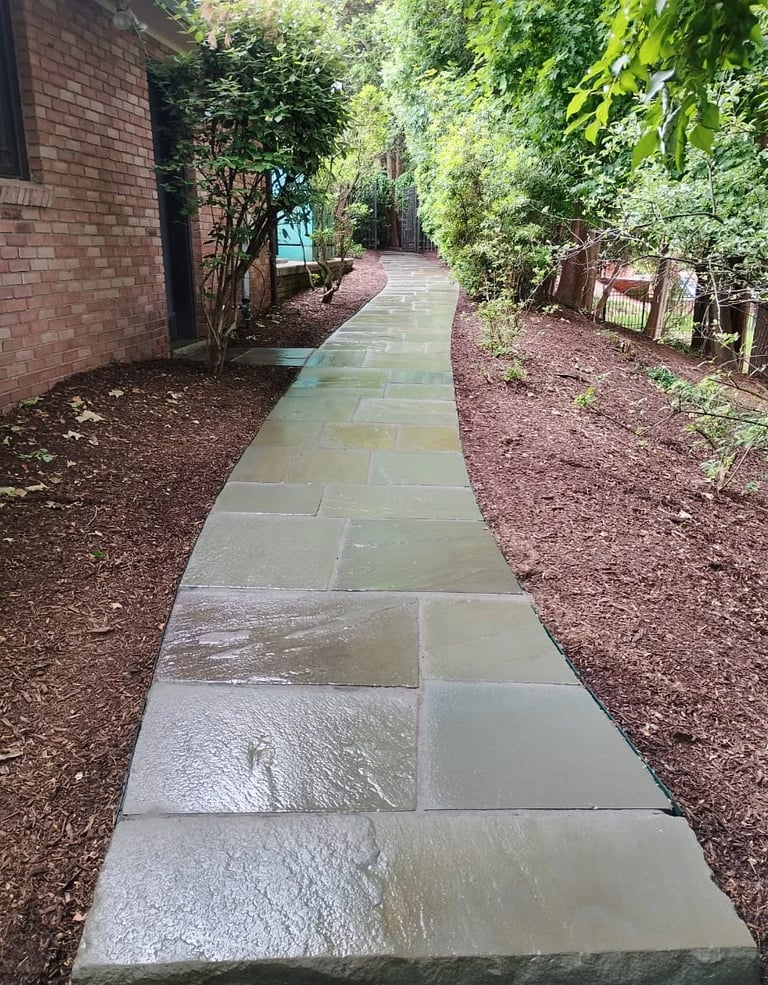 A paved stone pathway winds through a lush garden with green vegetation and trees on both sides, leading toward a brick building