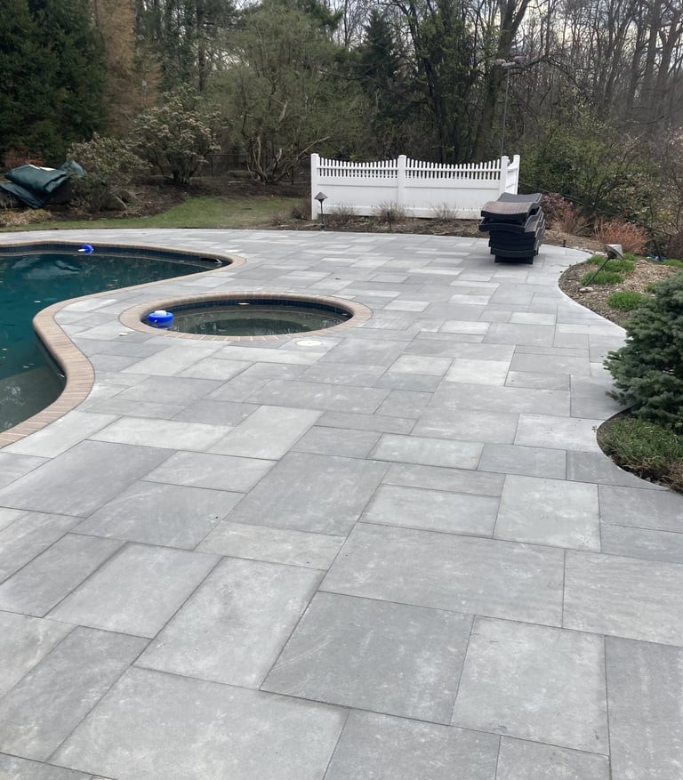 Backyard patio with pool and hot tub surrounded by stone pavers, white fence, and wooded landscape