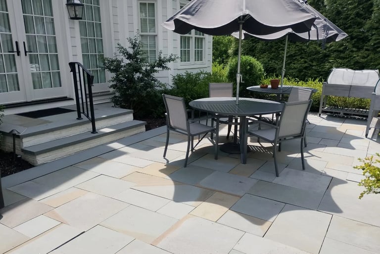 Modern patio with round black dining table, gray chairs, black umbrella, and potted plants on light stone pavers near white house