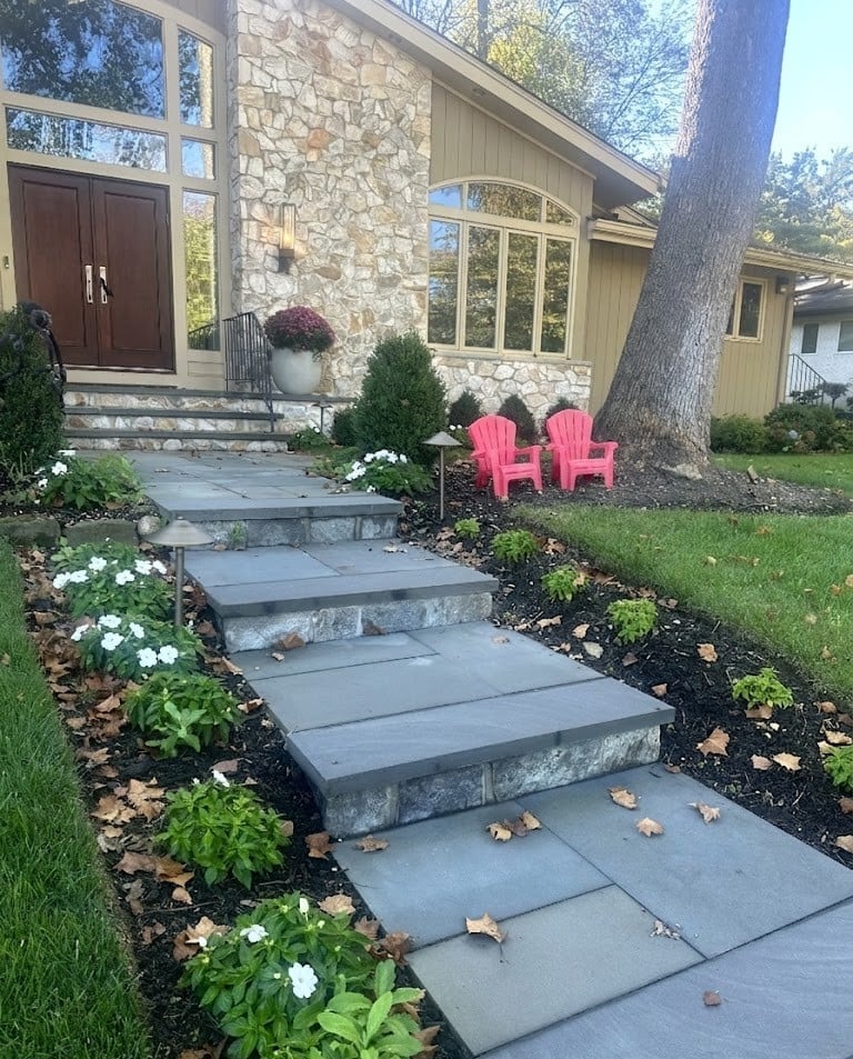 Modern home entrance with stone pathway, pink Adirondack chairs under a large tree, manicured landscaping with white flowers and shrubs