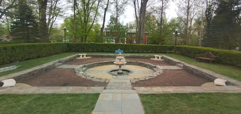 Formal garden with central fountain surrounded by geometric brick and stone pathways, hedges, and trees