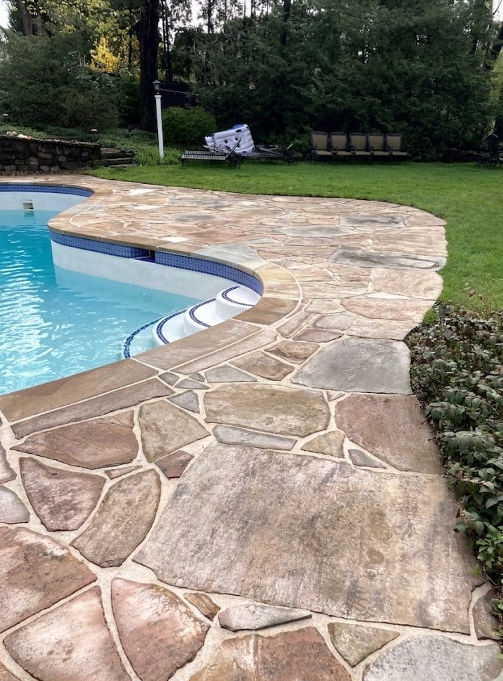 Backyard swimming pool with curved concrete patio and decorative pavers, surrounded by green lawn and tall trees