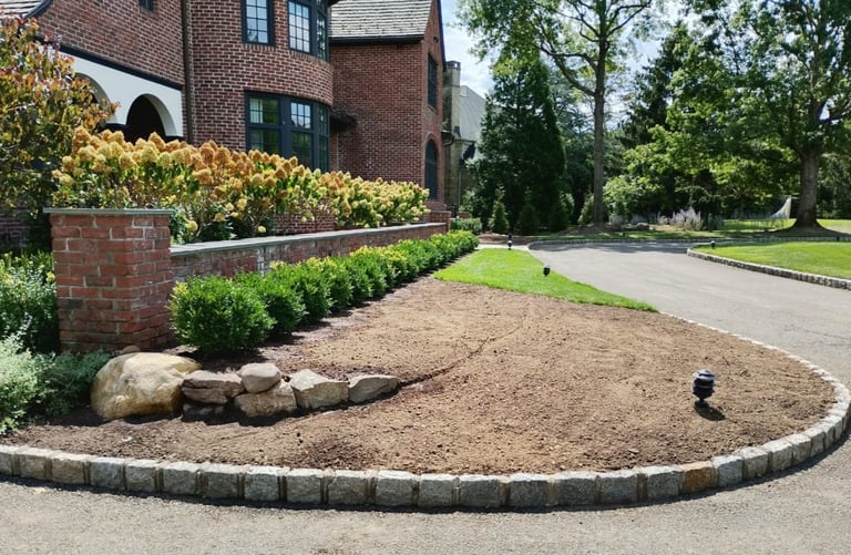 Curved brick garden bed with mulch and landscaping in front of brick house with tree-lined driveway