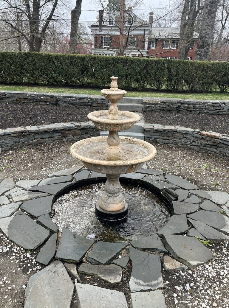 Three-tiered stone fountain in a circular slate basin, set in a garden with bare trees and a brick house in the background
