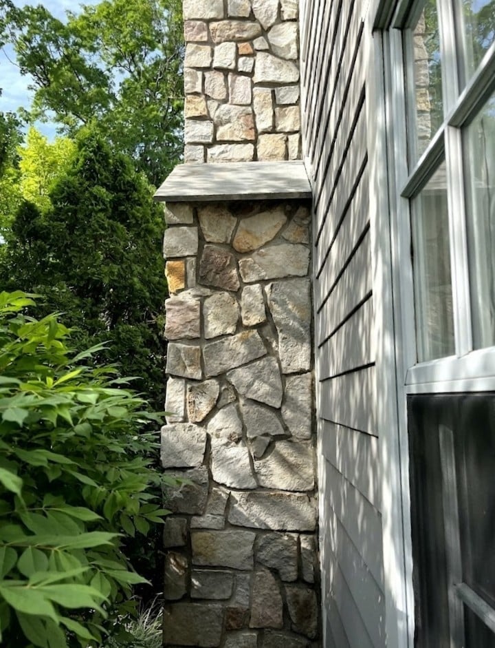 Close-up view of a stone chimney exterior against a house with windows and green foliage visible on the left