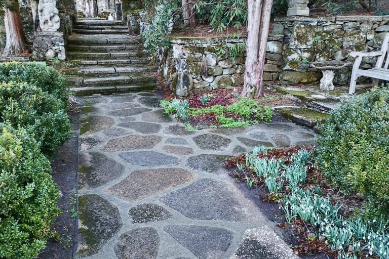 Scenic garden pathway with patterned stone tiles bordered by green shrubs and plants, leading through a wooded area with stone walls and steps