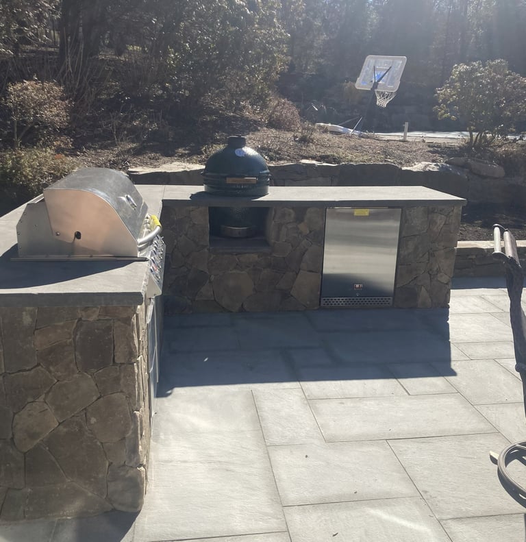 Outdoor kitchen setup with stone structure, dome grill, and refrigerator on concrete patio surrounded by trees