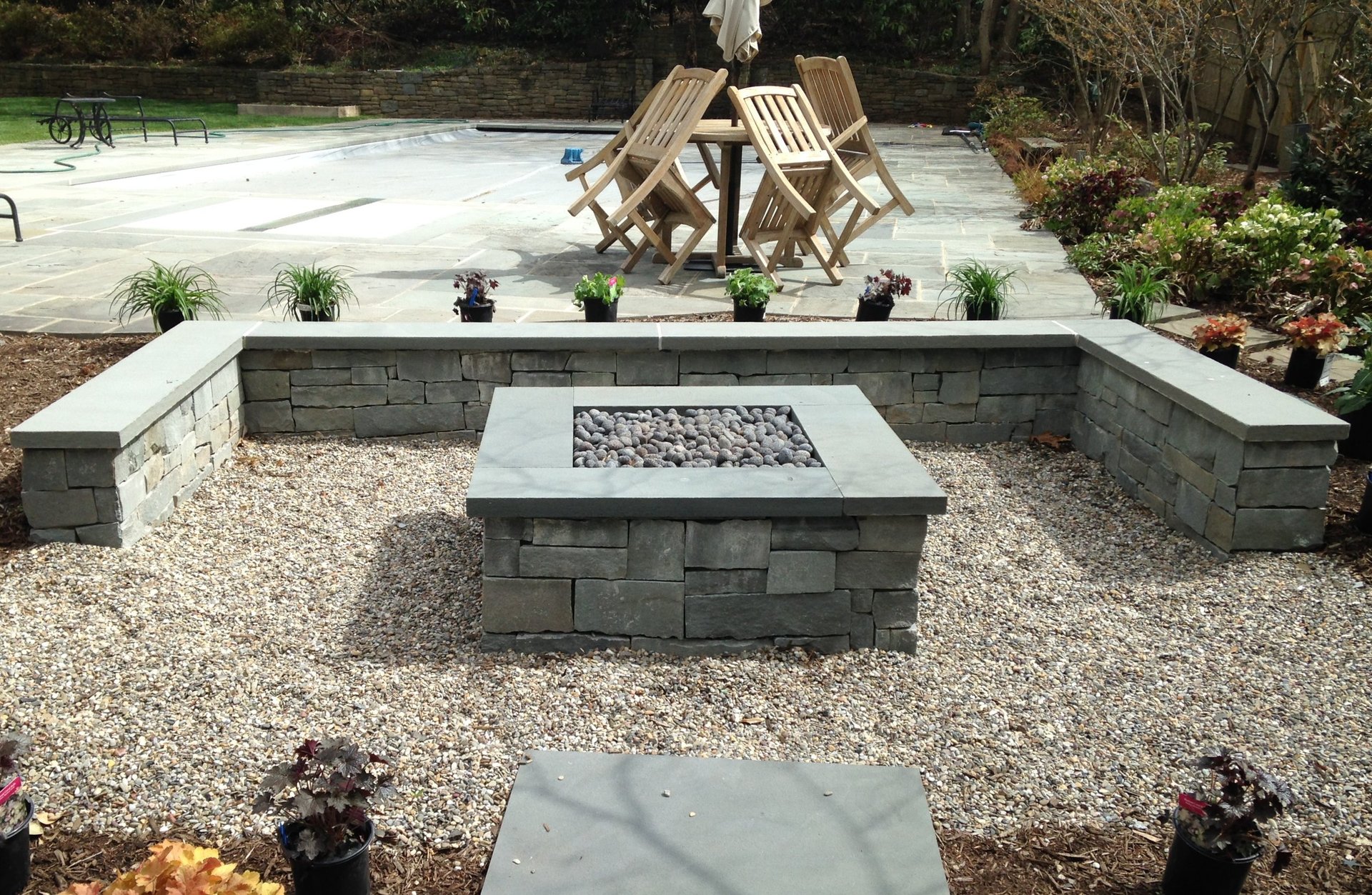 Backyard patio with built-in stone seating around a central fire pit, gravel flooring, wooden lounge chairs, and landscaping in the background