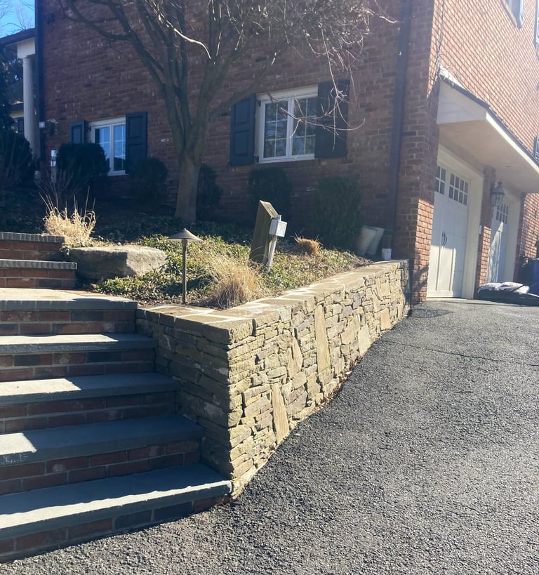 Stone retaining wall along a residential driveway with brick house and steps visible