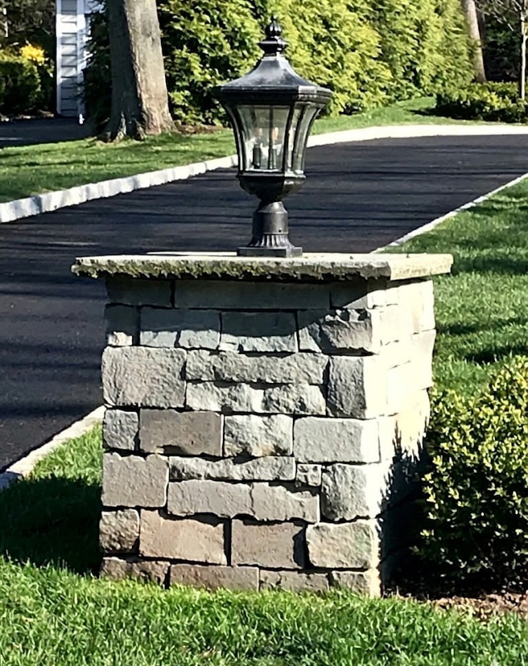 Stone column pillar with black lantern post light mounted on top, in residential driveway with trees and green lawn