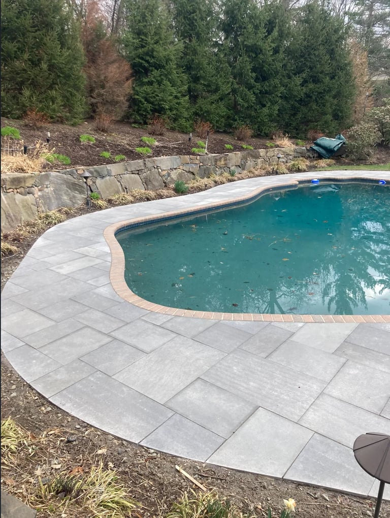 Backyard swimming pool with turquoise water, concrete patio, and stone retaining wall surrounded by tall evergreen trees