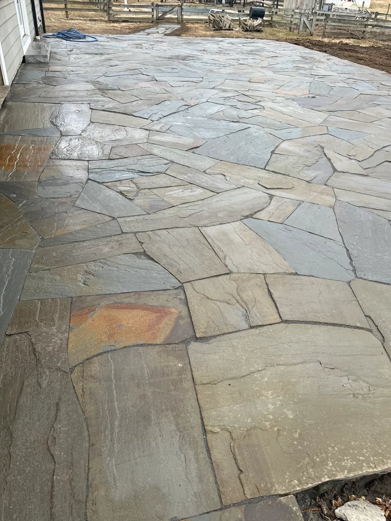 Irregular stone patio with various tan and gray flagstones laid in a random pattern, rural setting with fencing visible in background