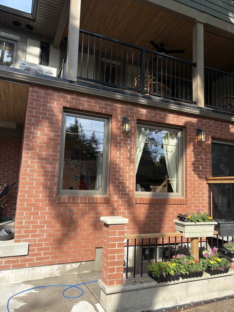 Brick facade of a two-story townhouse with large windows, metal railing on upper balcony, and planters with flowers at ground level
