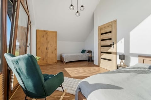 Modern bedroom with wooden flooring, teal armchair, white bedding, wooden doors, and pendant lights in a bright attic space