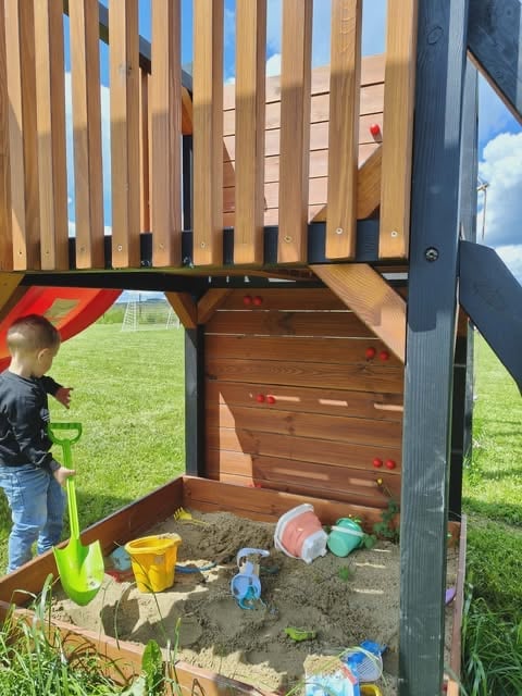 Young child playing in a sandbox under a wooden pergola structure in a backyard with grass and toys